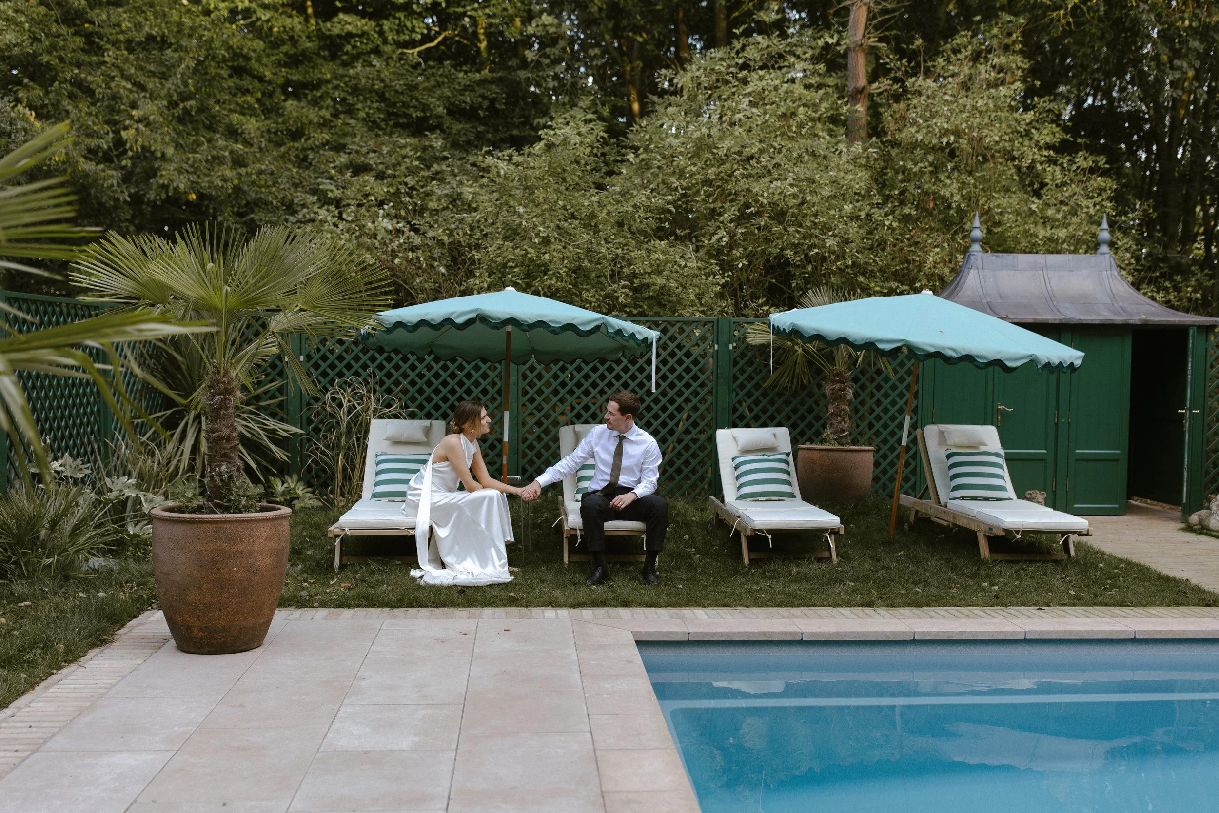 A couple in formal attire sitting by a poolside, holding hands, with sun loungers, umbrellas, large potted plants, and a small green garden shed in the background.