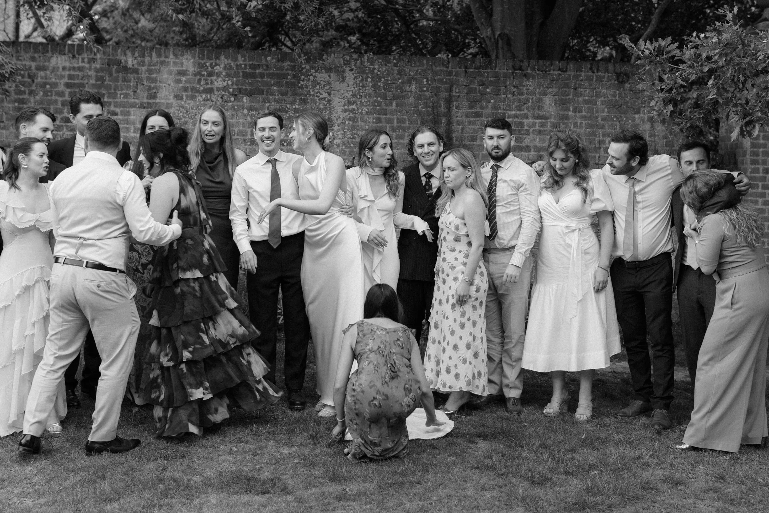 Group of people at an outdoor gathering or celebration, standing in front of a brick wall, engaging with each other, some smiling and some with neutral expressions.