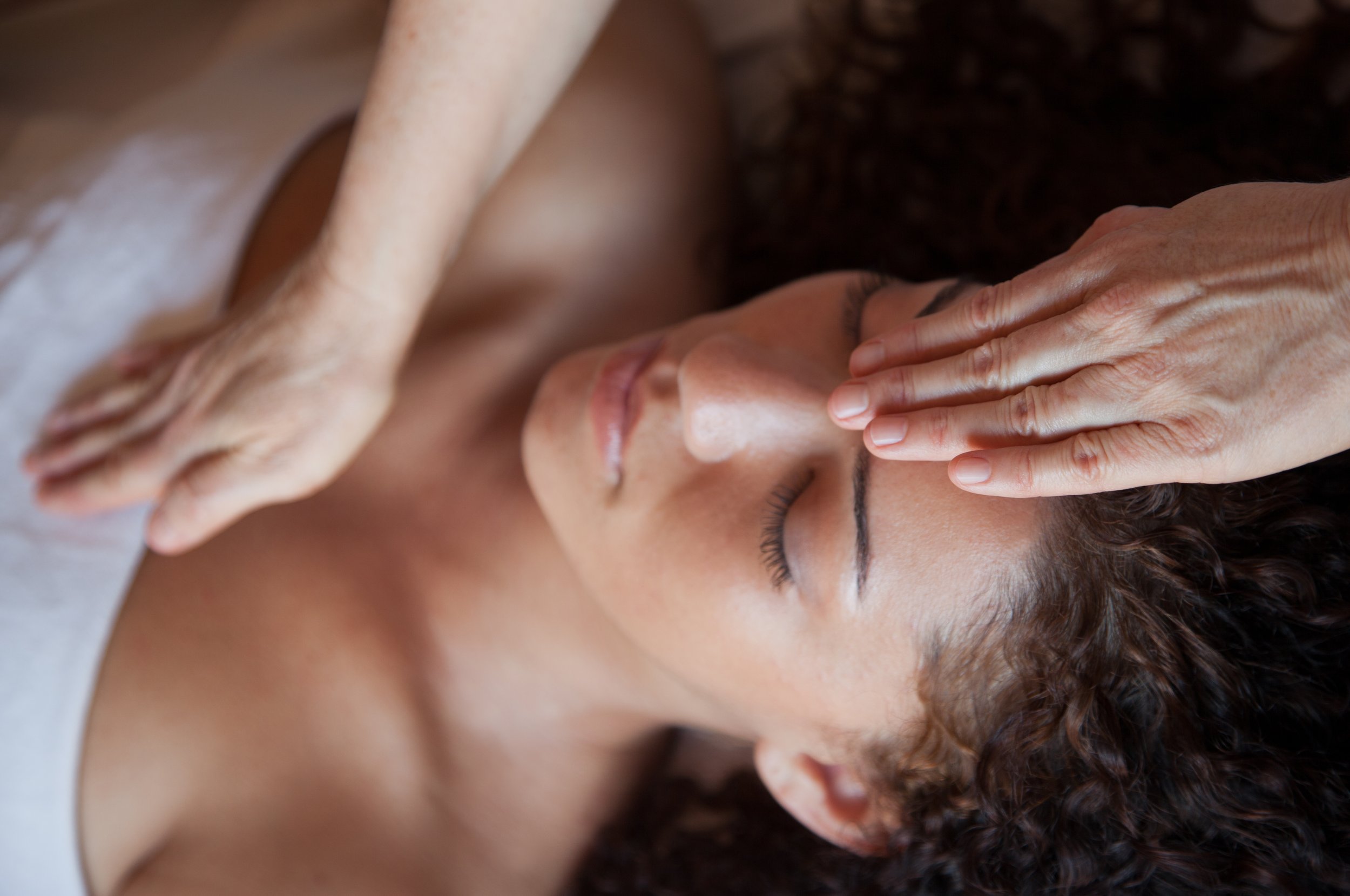 A woman receiving a facial massage, lying with eyes closed, with a therapist's hand on her forehead.