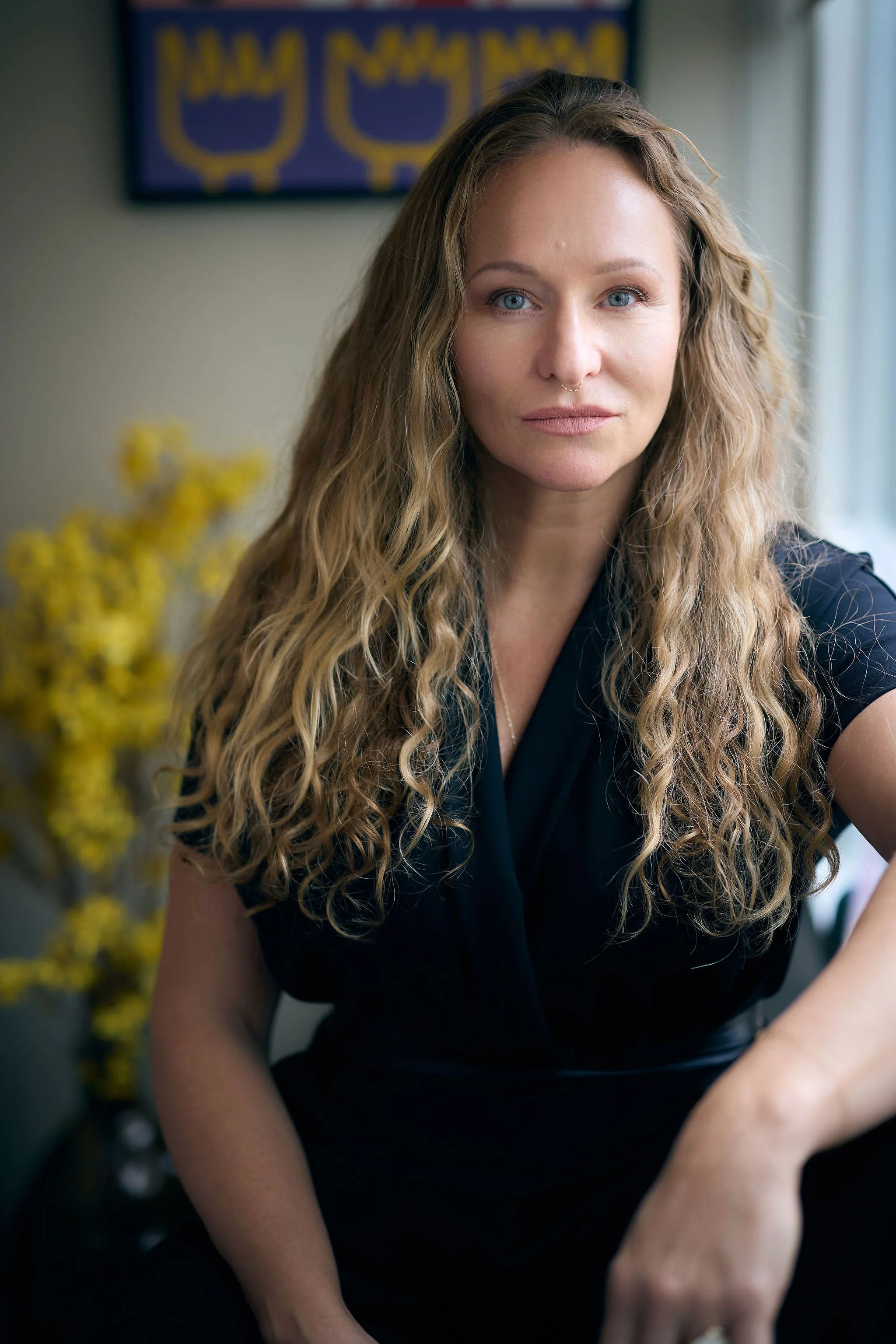 Close-up of a woman with long, wavy blond hair and blue eyes, wearing a black top, sitting indoors with a blurred background including yellow flowers and artwork on the wall.