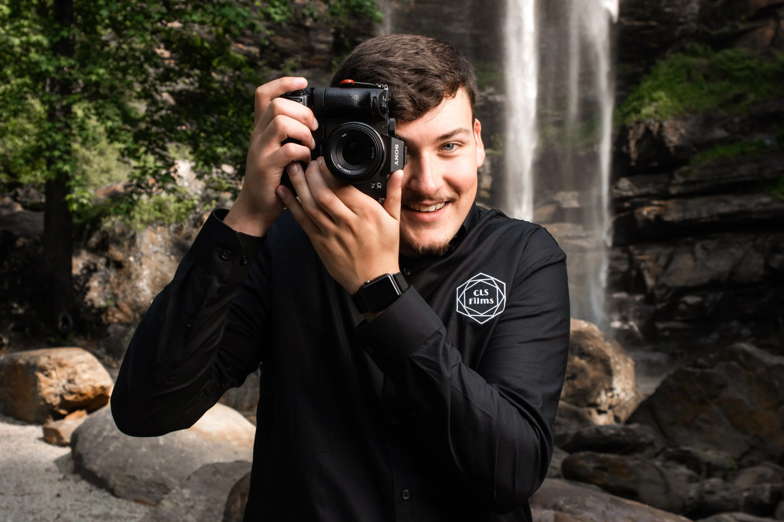 A young man with short brown hair and blue eyes holding a camera to his face, smiling, in front of a waterfall surrounded by greenery and rocks.