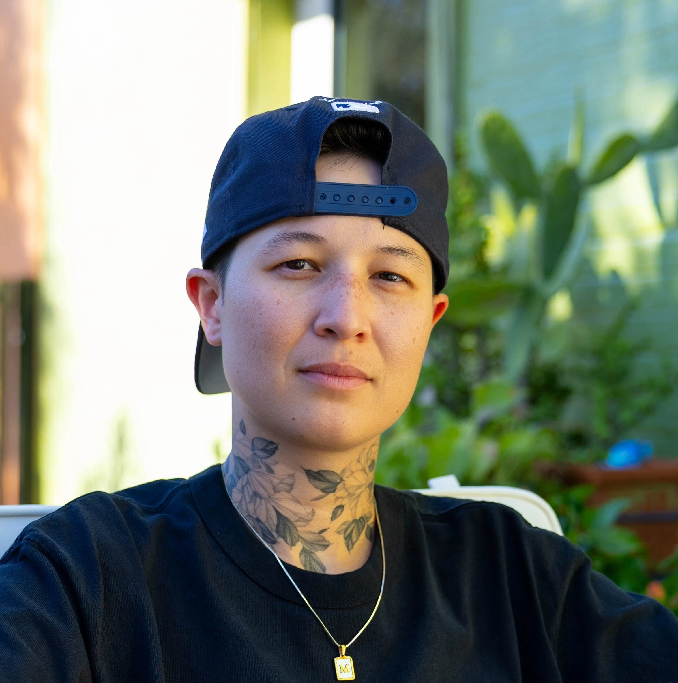 A person with short dark hair, freckles, and a floral tattoo on their neck, wearing a gray sweatshirt, standing against a dark textured wall.