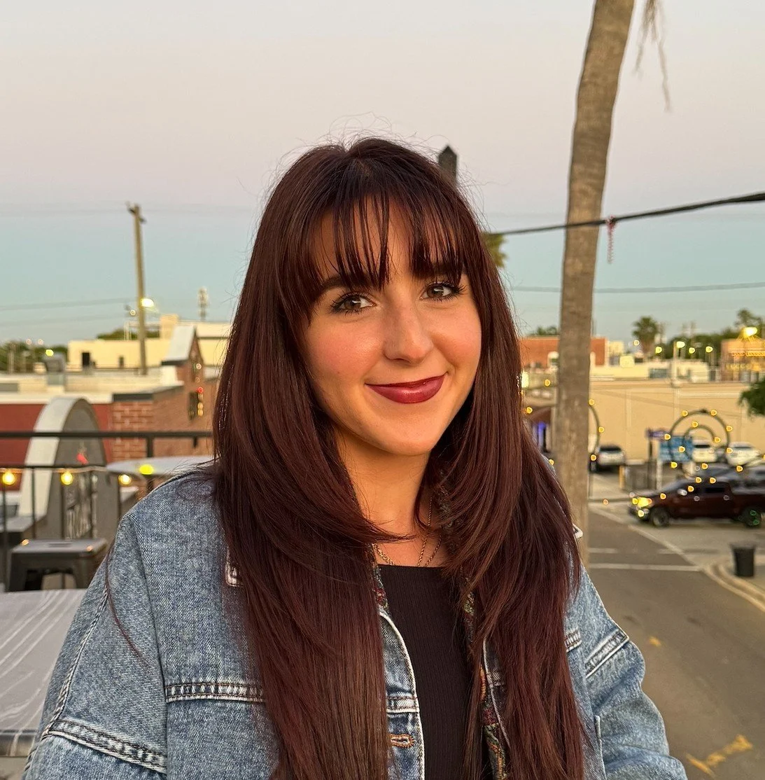 A woman with long red hair and bangs smiling outdoors, wearing a denim jacket and black top, with a parking lot and palm trees in the background during sunset.