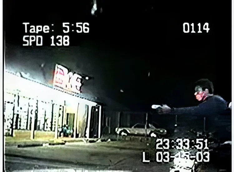 Nighttime scene of a police dashcam showing a woman with glasses wearing a mask and dark clothing, pointing a gun. The background features parked cars and a building, and there are on-screen data including time, speed, and date.