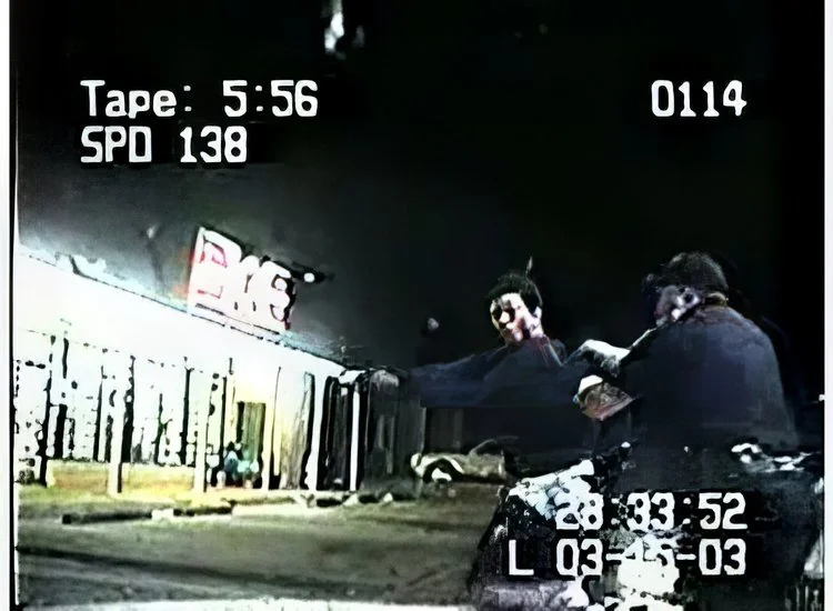 Nighttime scene showing two women, one with curly hair and another with straight hair, standing near a car, with one woman wearing gloves, beside a building with a lit sign and a police vehicle nearby.