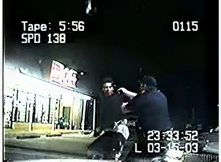 A police officer aiming a firearm at a person outside a Taco Bell restaurant at night. The scene is captured by a dashcam, with a timestamp showing 11:33 PM on March 15, 2003.