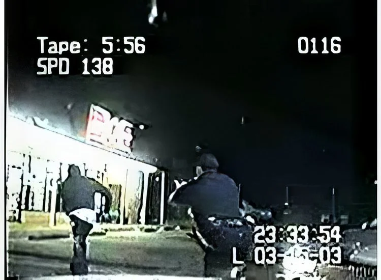 Nighttime police dashcam footage of two officers on bicycles near a building with a lit sign, seen from behind. Timestamp indicates 23:33:54.