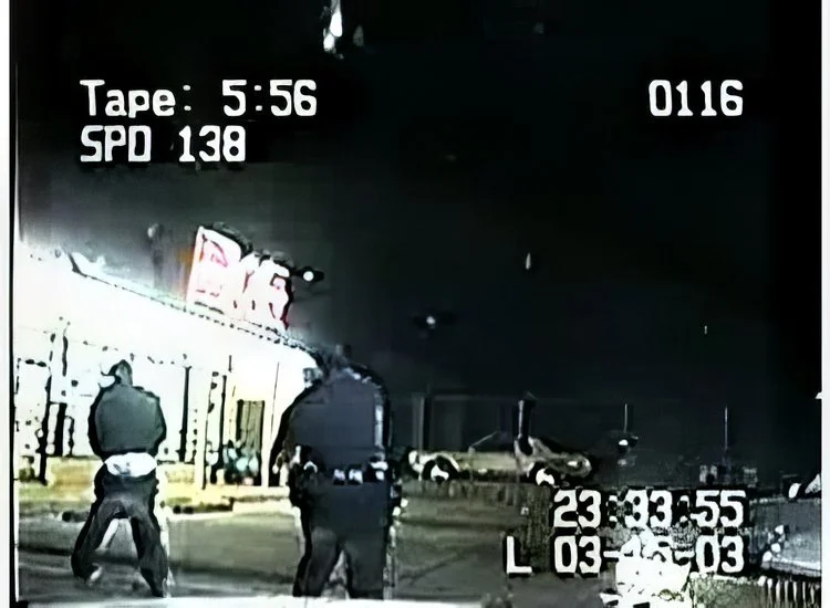 Nighttime police dashboard camera image showing two police officers standing in front of a vehicle with a police car nearby. The image includes on-screen data such as tape time 5:56, SPD 138, and date 03-20-03.