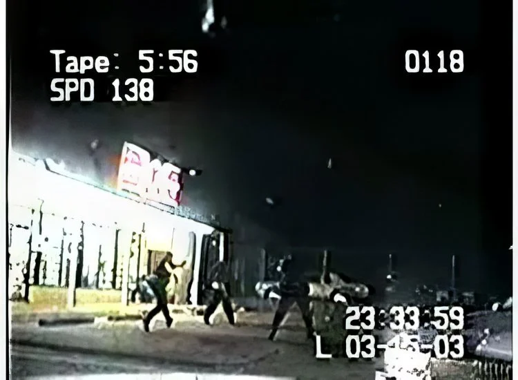 Nighttime scene showing a group of people walking on a sidewalk in front of a store. Several cars parked along the street. A large illuminated sign with partially visible text is above the store entrance. On-screen data displays a timer, date, and other numbers.