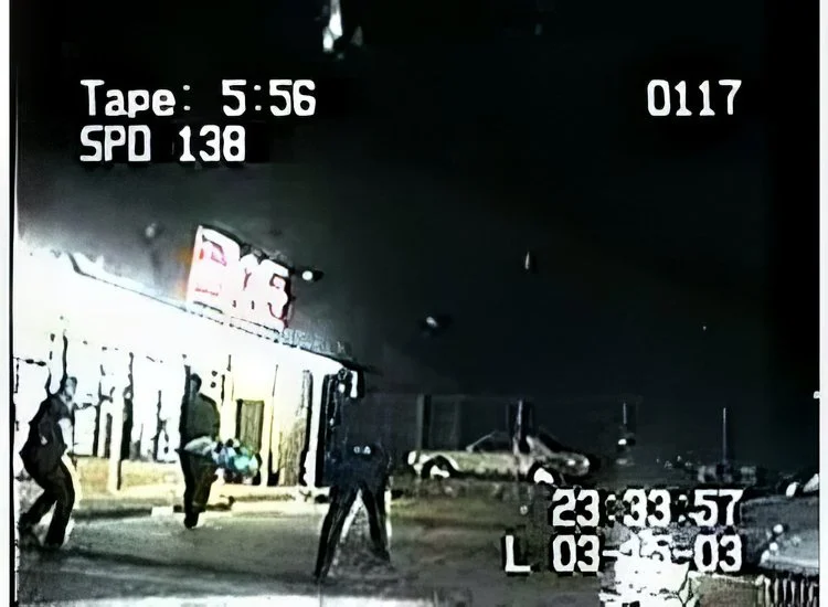 A nighttime scene of a parking lot with several people walking, some carrying items, near a building with a sign, and parked cars in the background.