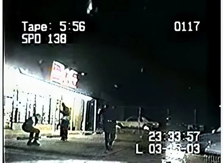Nighttime street scene with three pedestrians walking past a graffiti-covered wall, and a blurred image of a helicopter in the background. CCTV footage timestamp shows 23:33:57.