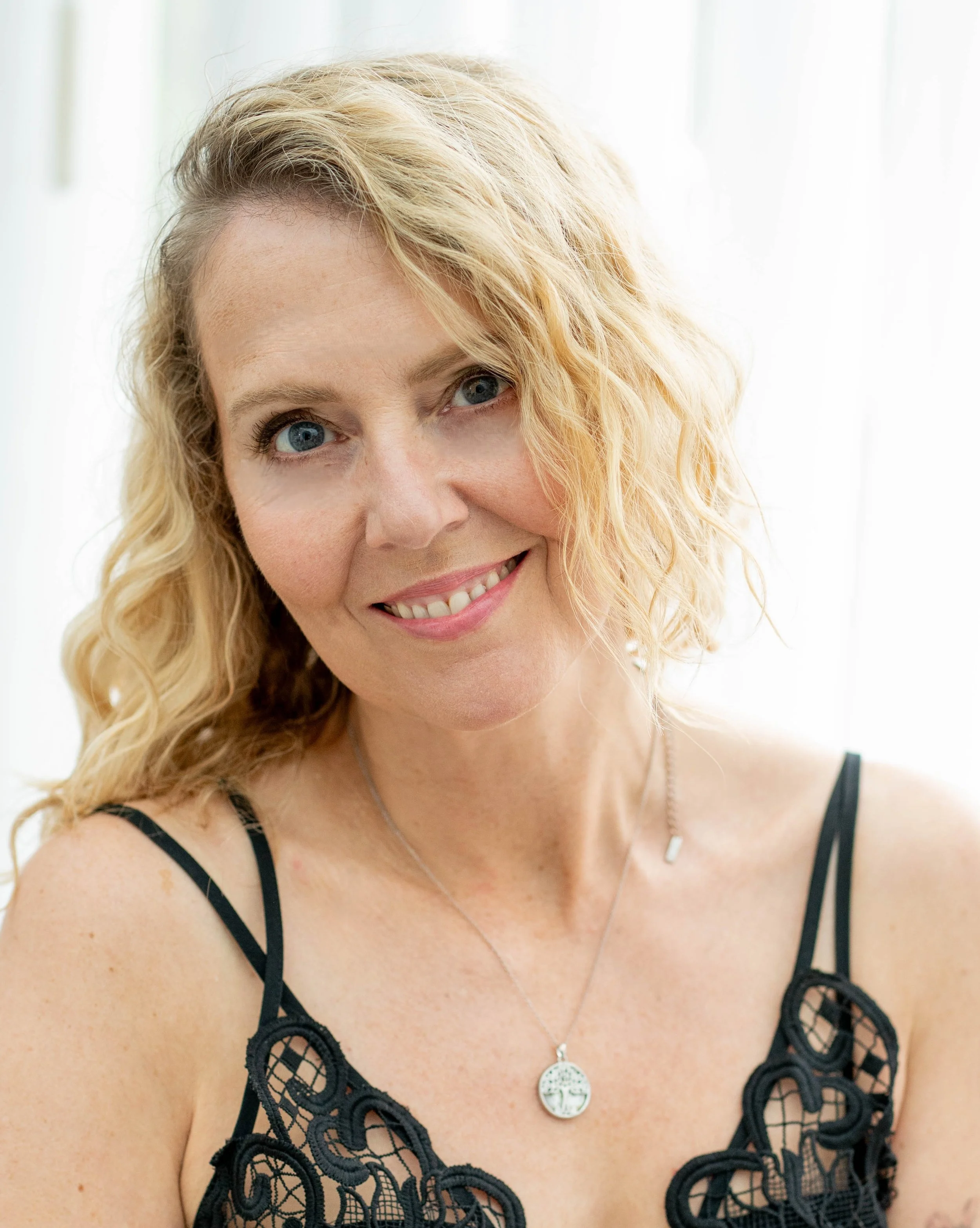 Satya Tremaine somatic therapist singer songwriter smiling, with curly blonde hair, wearing a black lace dress