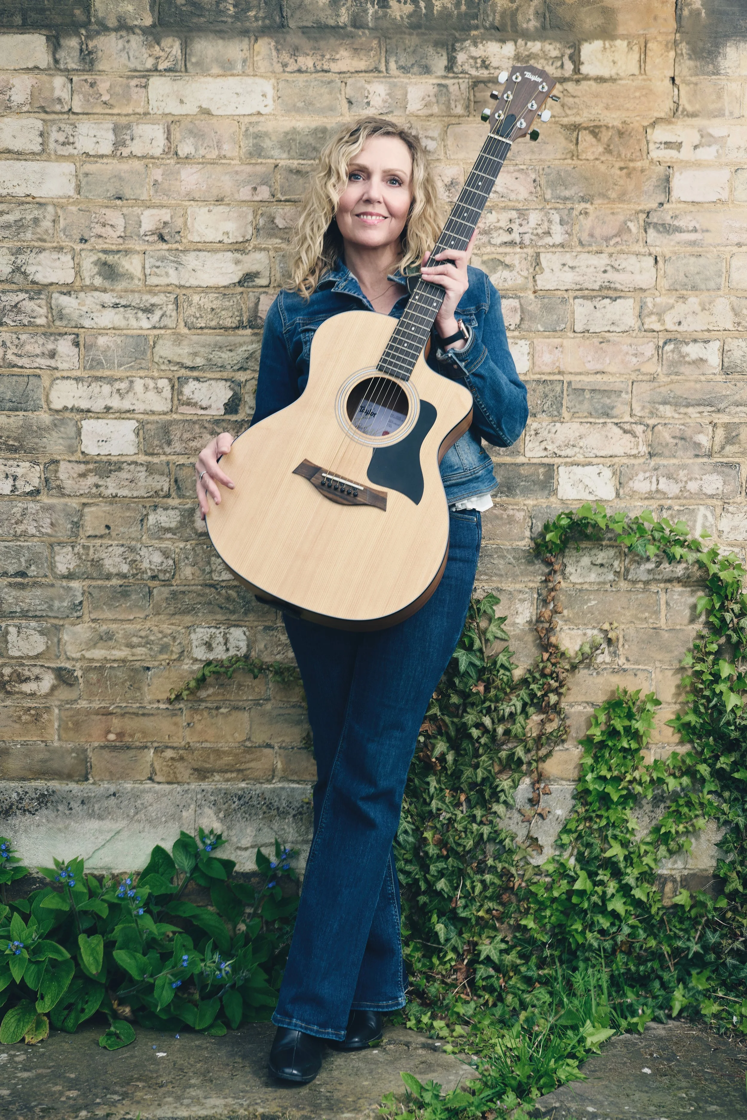 Satya Tremaine somatic therapist singer songwriter holding an acoustic guitar, with a brick wall and green plants in the background.