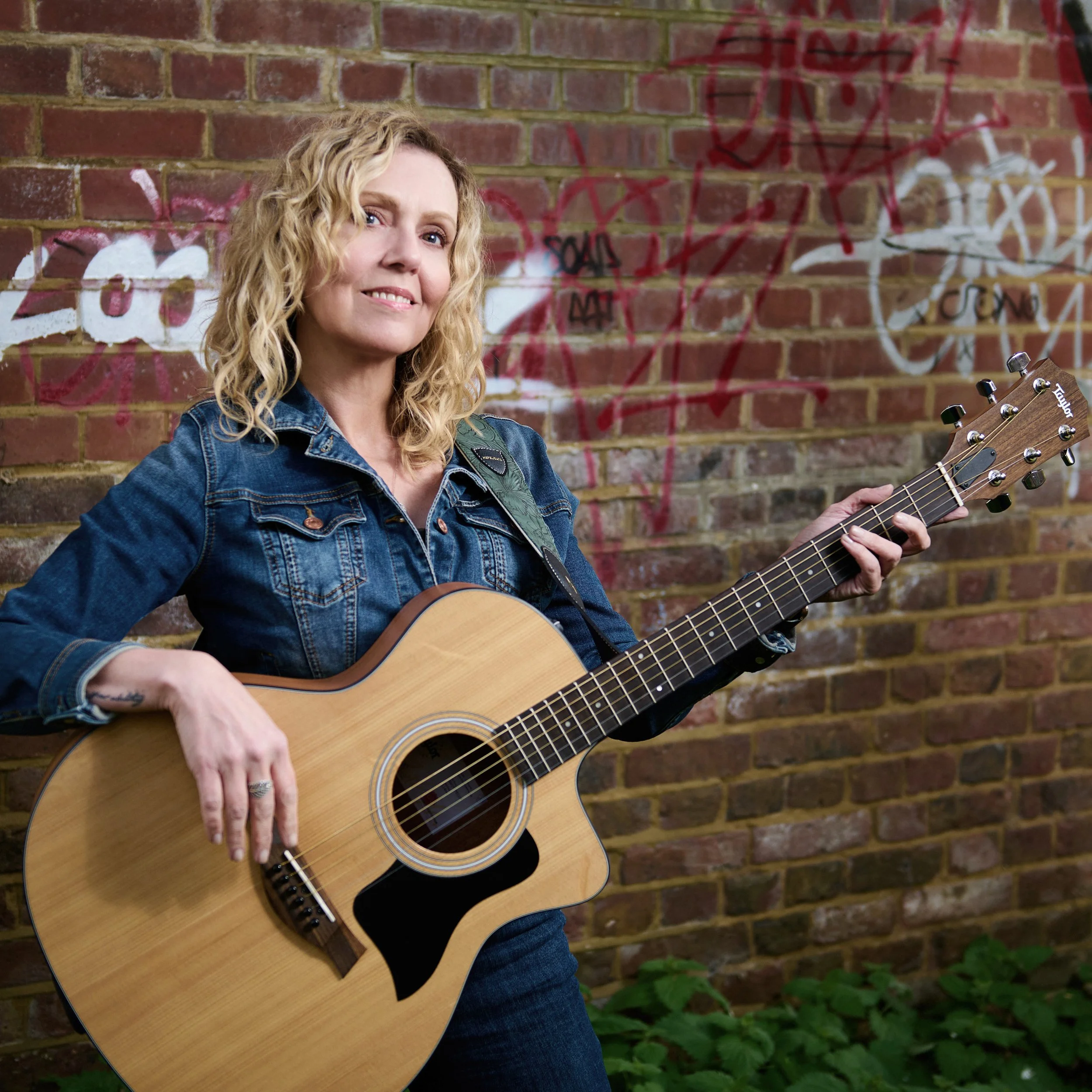 Satya Tremaine somatic therapist singer songwriter smiling while holding an acoustic guitar, standing in front of a brick wall with graffiti and some greenery.