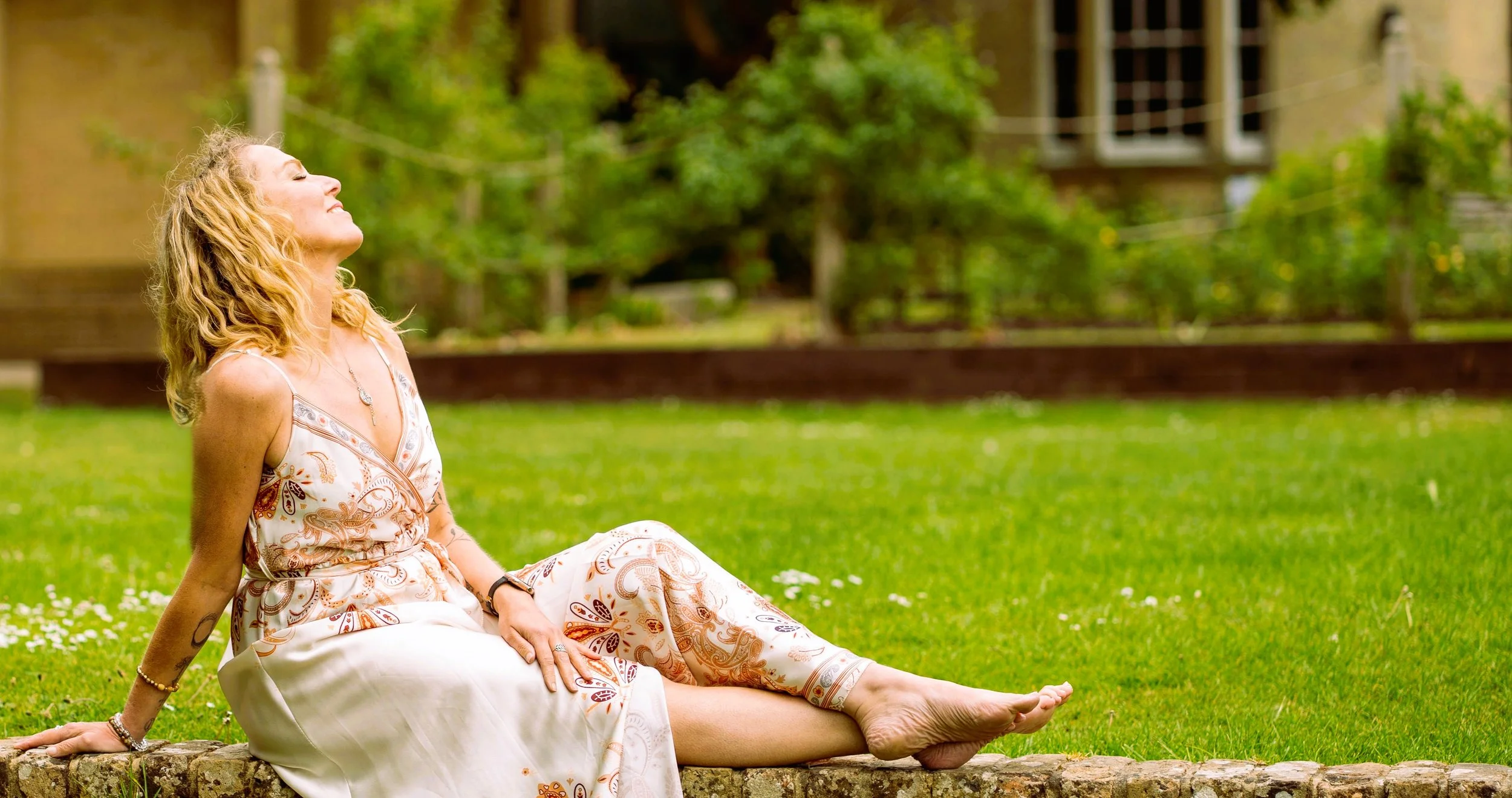 Satya Tremaine somatic therapist/ singer songwriter sitting on a stone border in a grassy field, smiling and looking up, wearing a patterned summer dress.