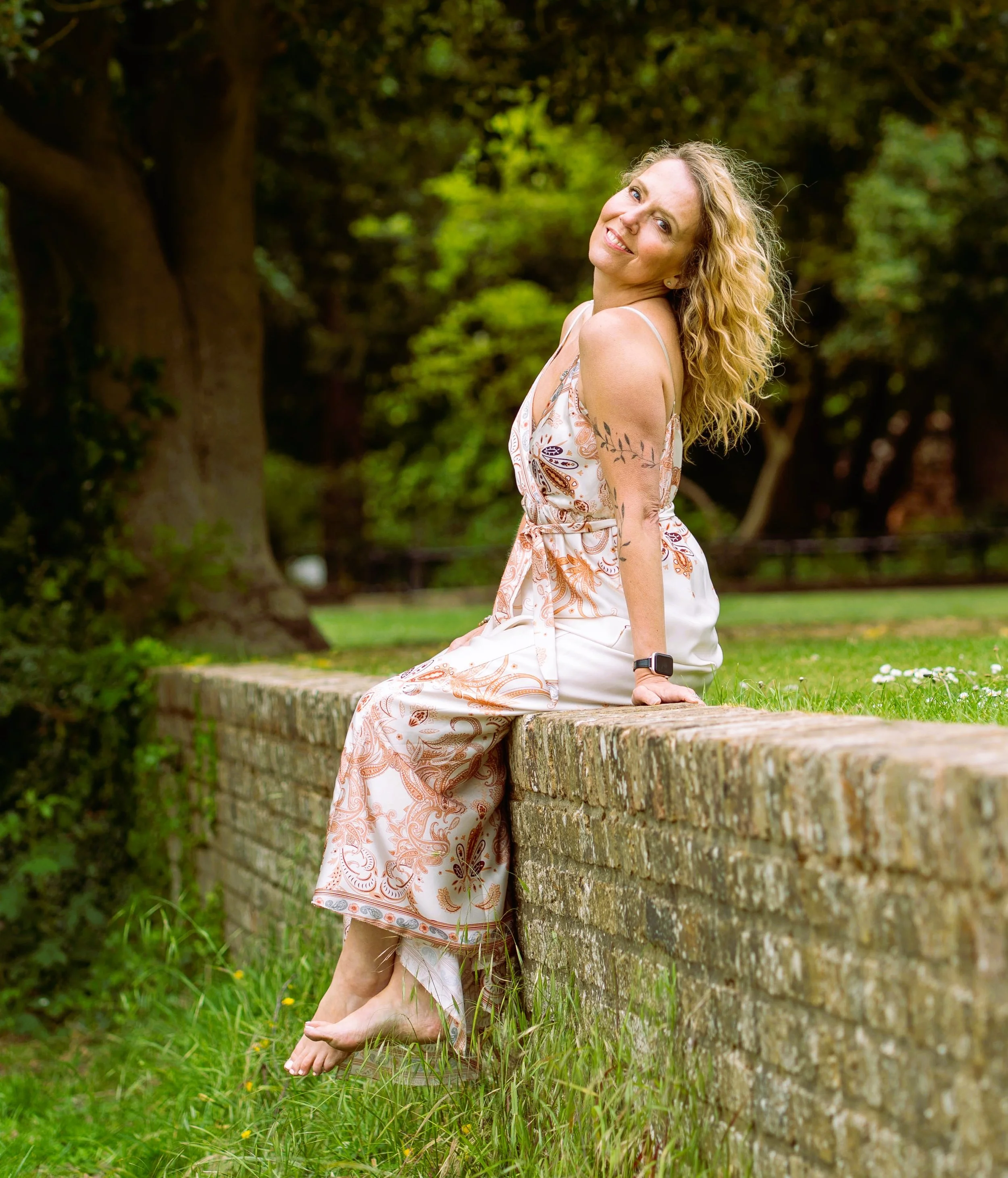 Satya Tremaine somatic therapist singer songwriter smiling and looking at the camera, wearing a white patterned dress, with trees and green grass in the background.