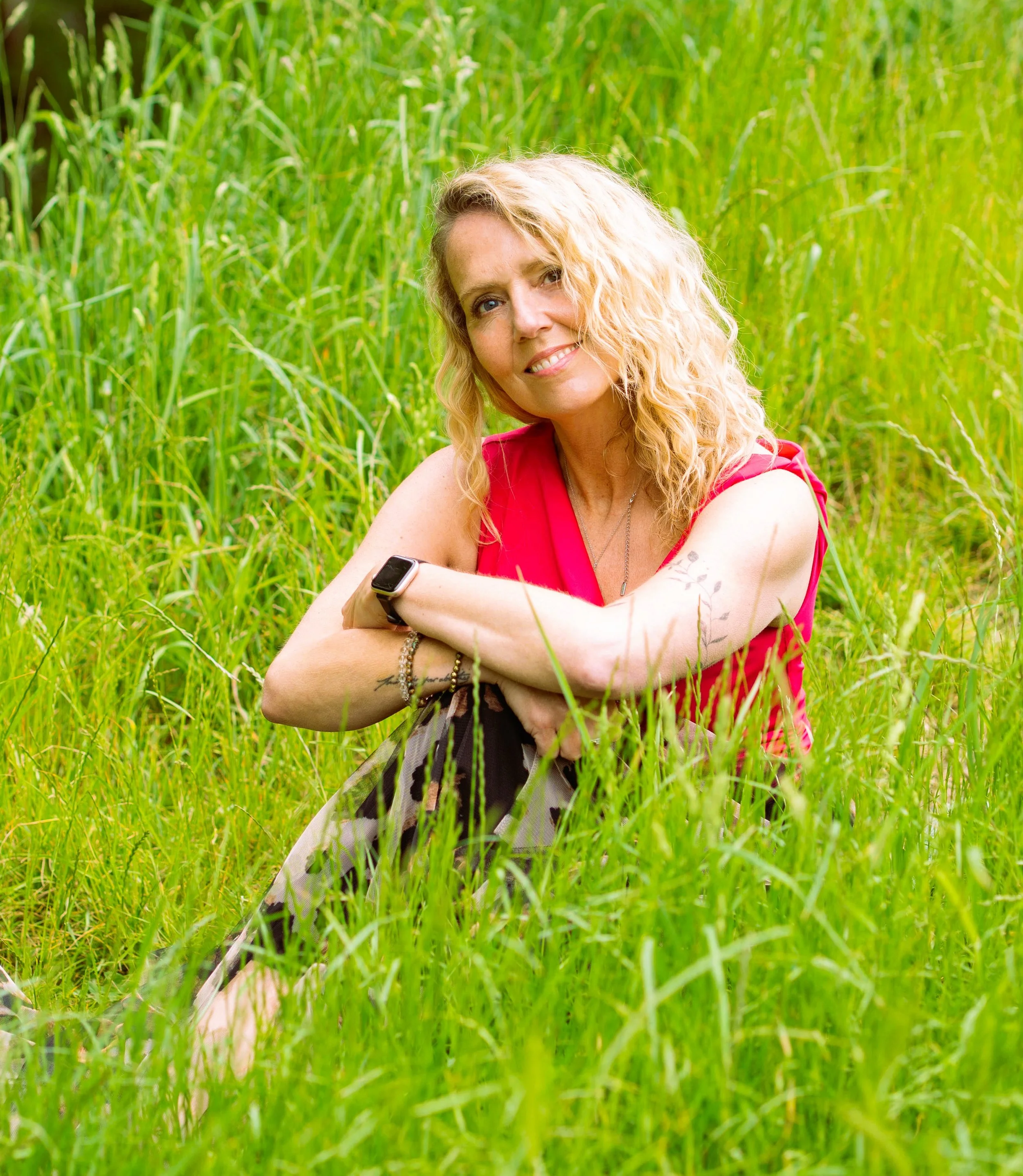 Satya Tremaine somatic therapist/ singer songwriter sitting on a stone border in a grassy field, smiling.