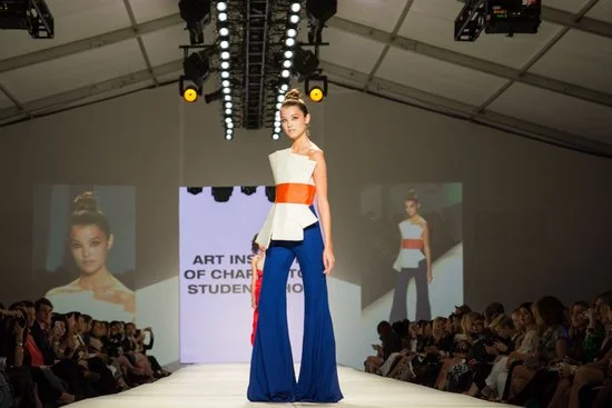 A model walks the runway in a fashion show wearing a white, orange, and blue outfit, with an audience seated on either side. The backdrop displays text related to the Art Institute of Charlotte's student show.