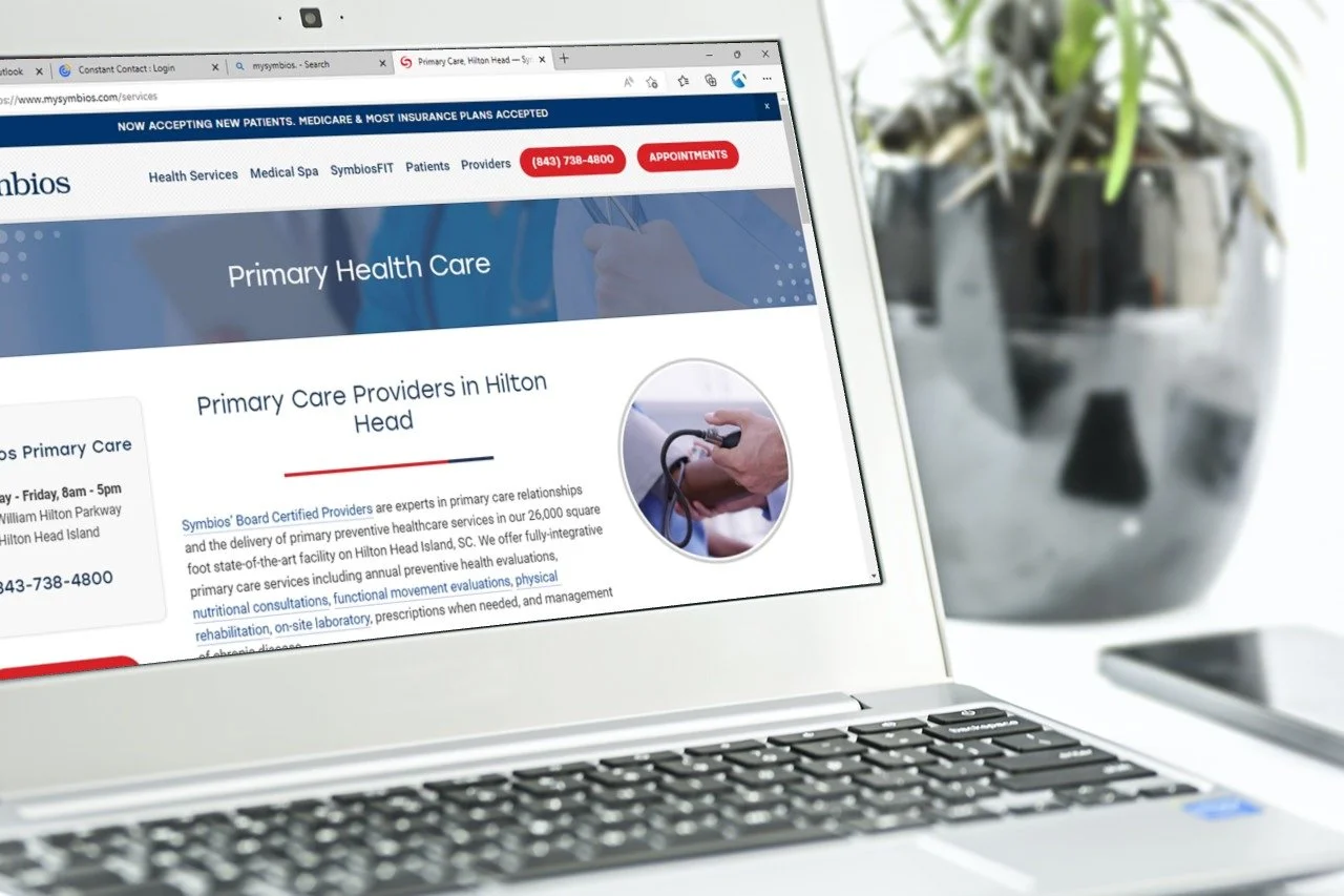 Laptop displaying a healthcare website for "Primary Care Providers in Hilton Head" with navigation links, appointment button, and plant in the background.