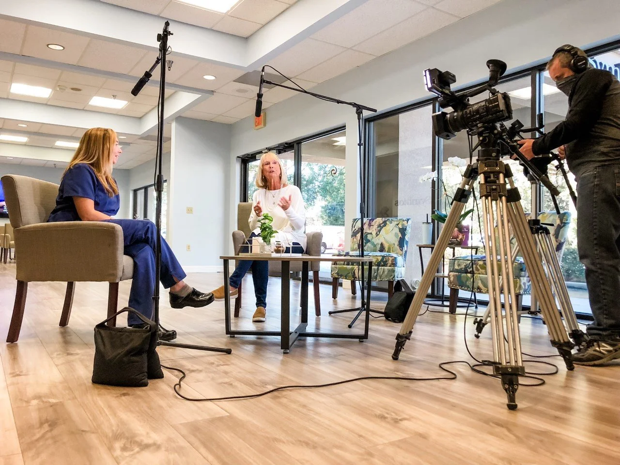 A medical professional in blue scrubs and another person are seated in chairs during an interview setup
