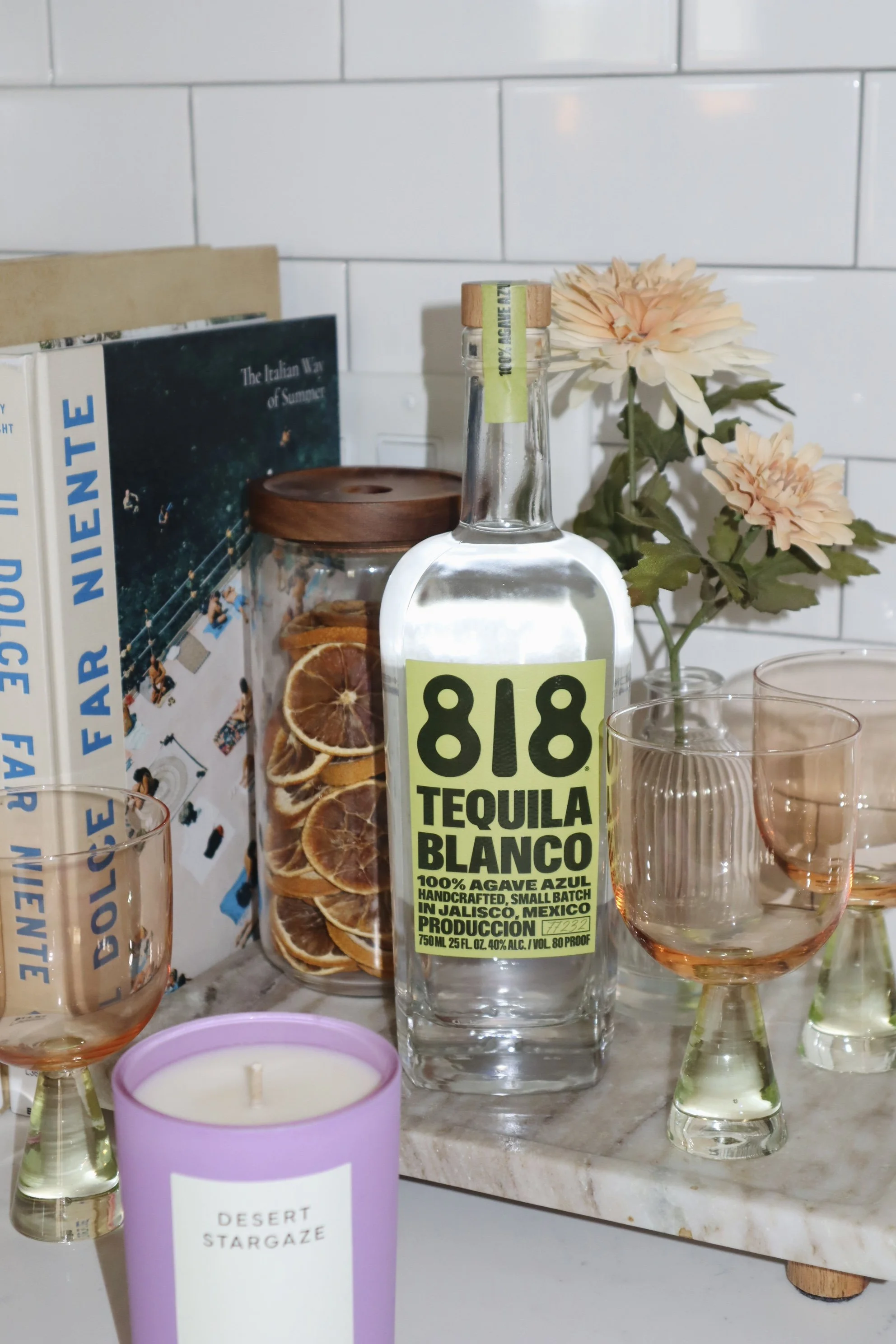 A clear bottle of blanco tequila with a yellow label, pink glasses, a jar of dried orange slices, a potted flower, a purple candle, and books on a marble countertop against a white tiled wall.