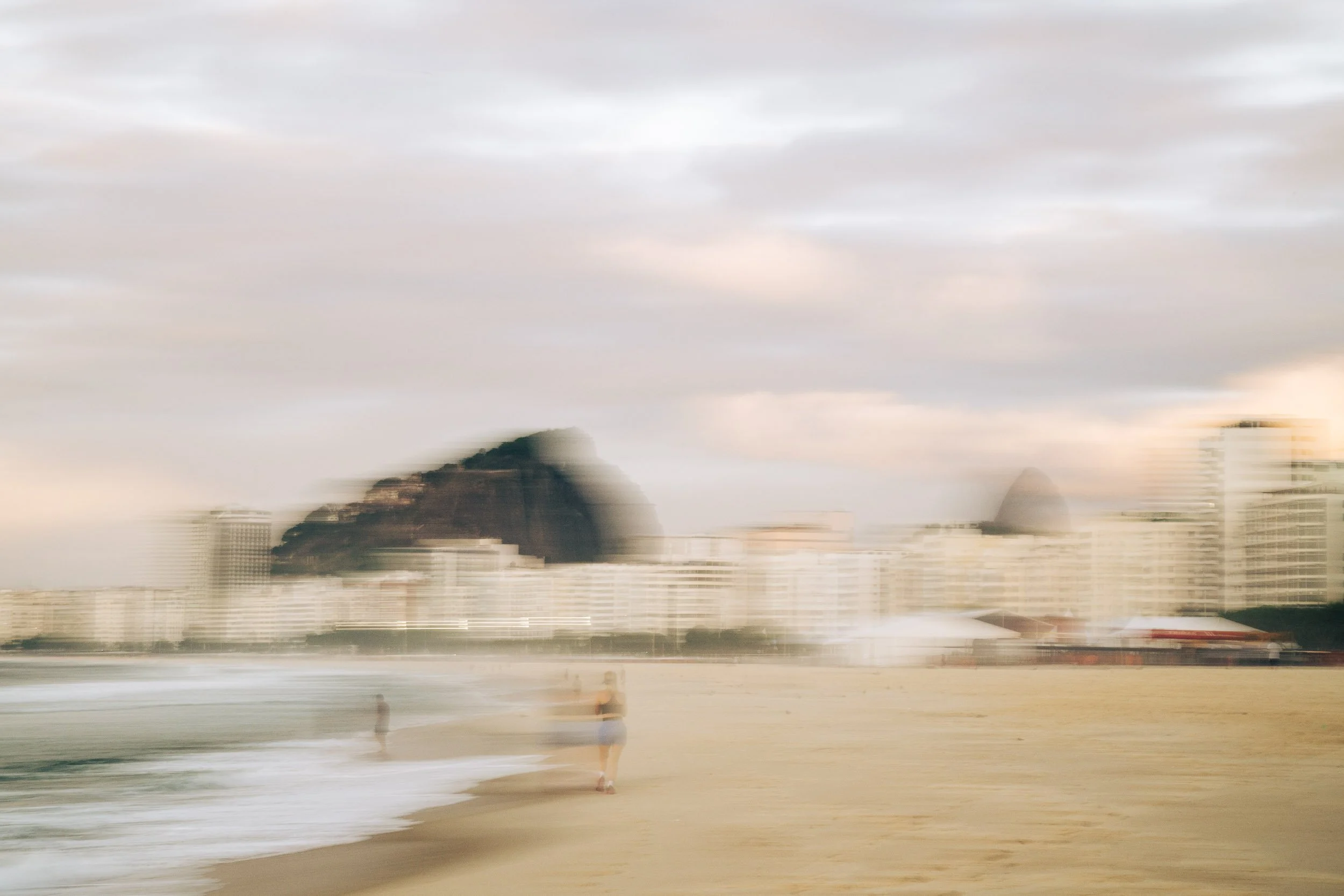 Motion-blurred view of Rio’s beachfront and city under bright daylight, with softened buildings, shoreline, and figures conveying heat, movement, and rhythm.