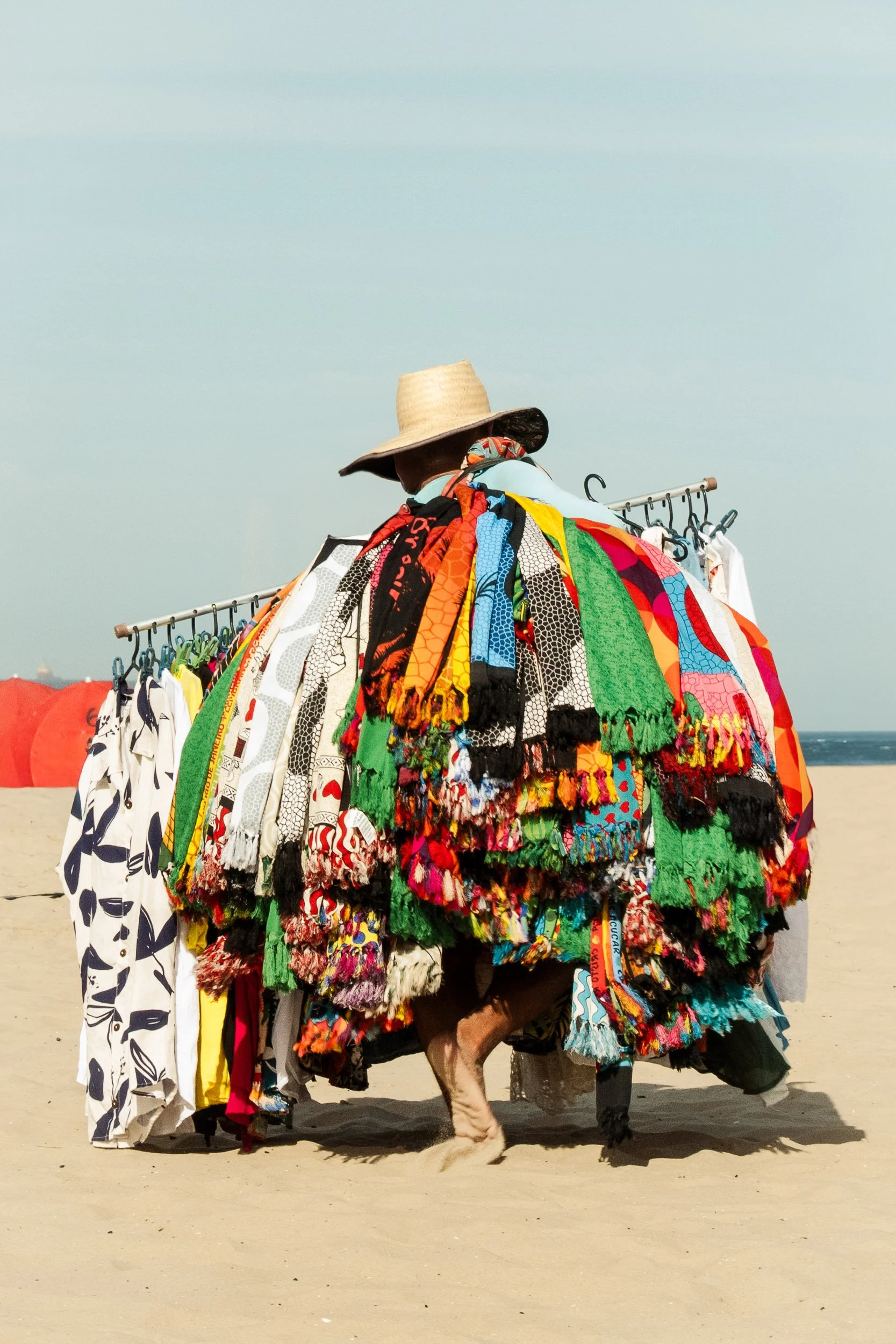 A barefoot fabric seller walking across sand in Rio, carrying a rack of brightly coloured fabrics against a pale sky and shoreline.
