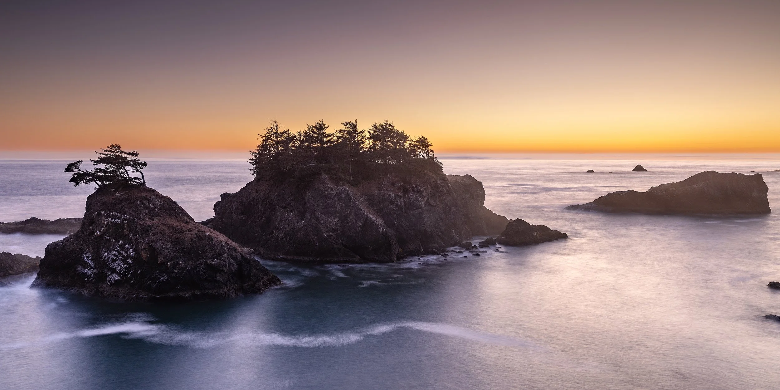 A seascape at sunset with rocky islands and trees, calm water, and a colorful sky.