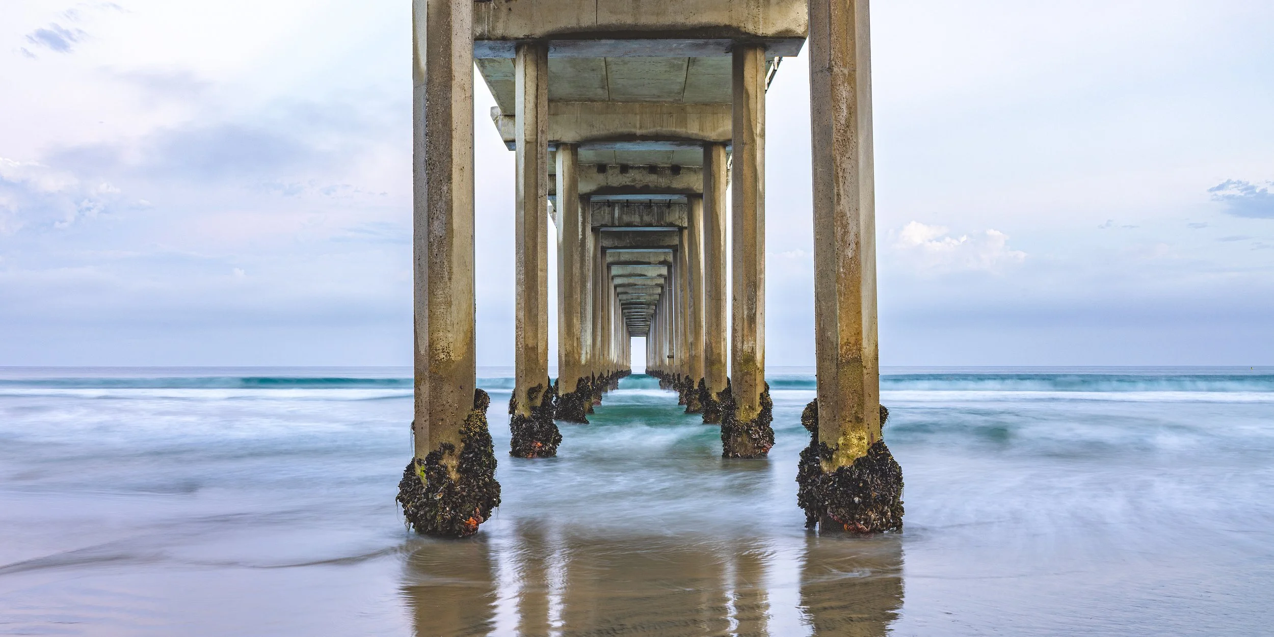 View of a concrete pier extending into the ocean, with moss-covered supports at low tide, overcast sky