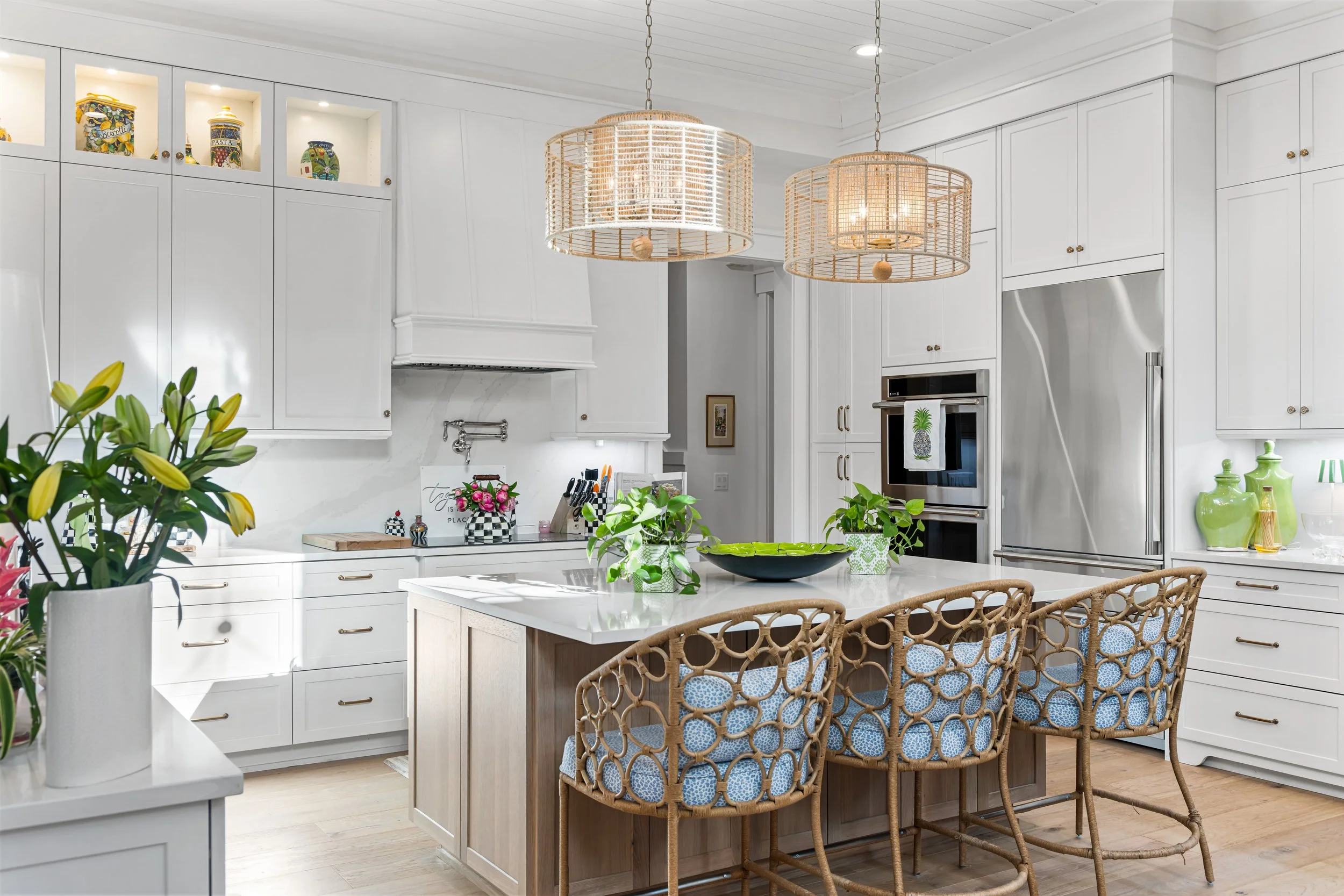 Bright white kitchen with a central island, wicker chairs with blue cushions, and potted plants. Open shelving with decorative jars, and stainless steel refrigerator and double oven.