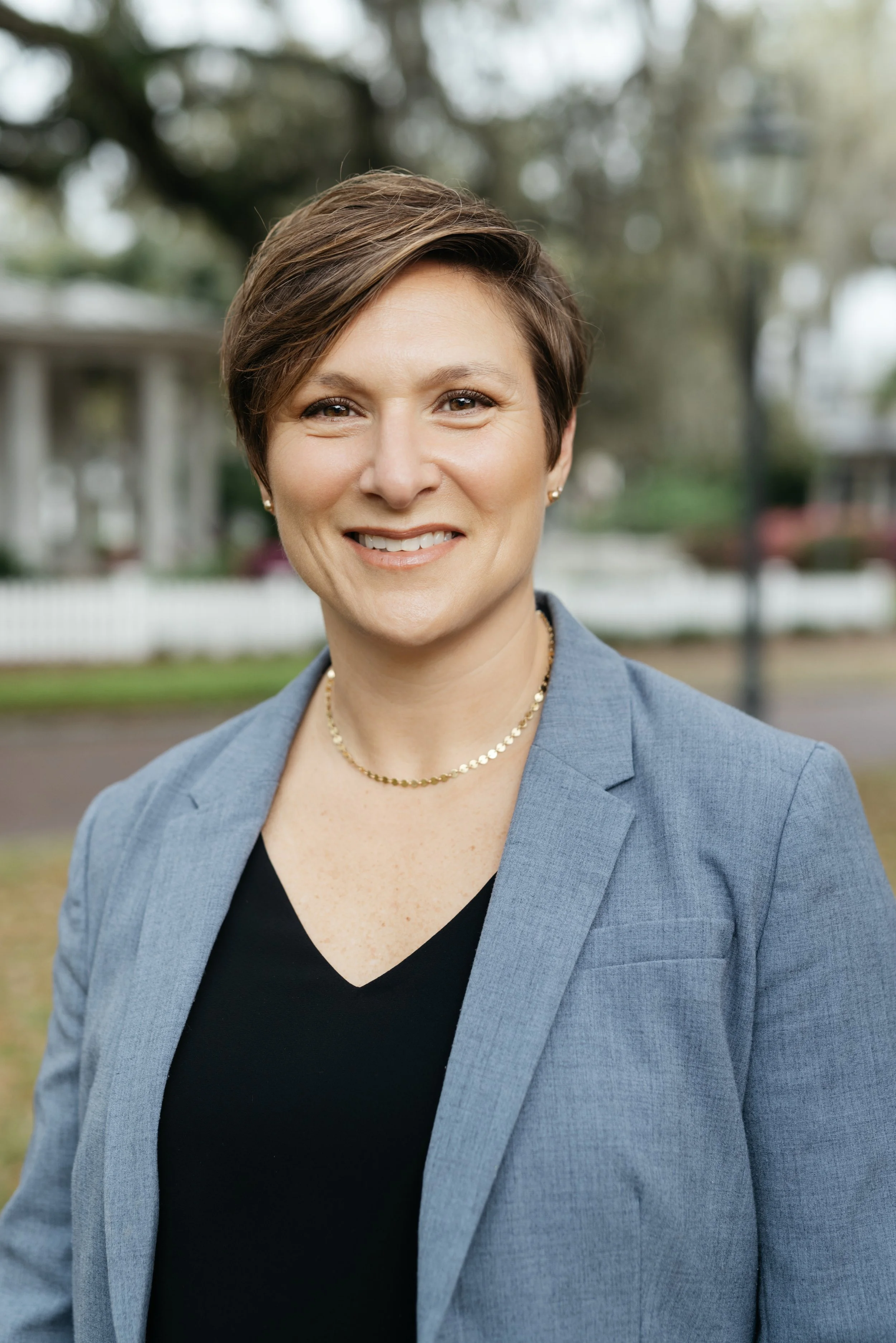 A woman with short brown hair smiling, wearing a gray blazer, black top, and a gold necklace, outdoors with blurred trees and houses in the background.
