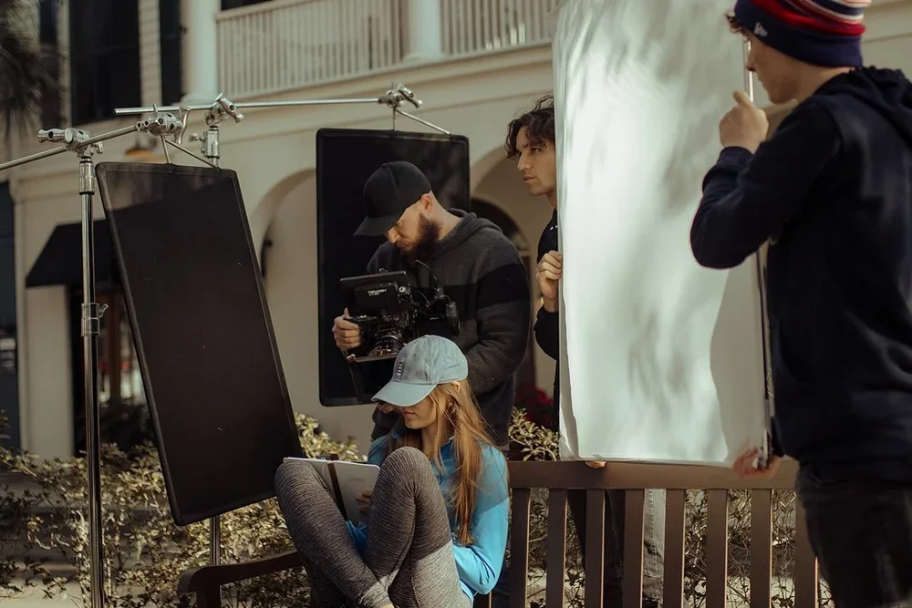Filmmakers filming a young woman sitting on a park bench with a notebook, surrounded by filming equipment and crew.