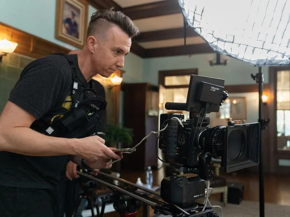 A filmmaker looking at a professional camera setup on a tripod, inside a cozy room with wooden wall paneling, paintings, and a large softbox light overhead.