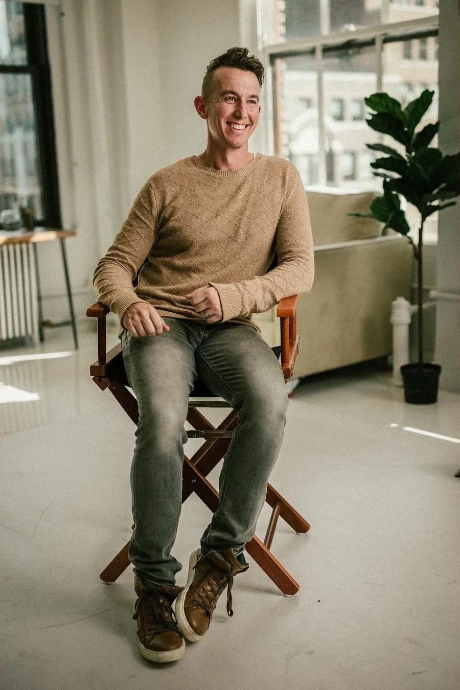 A smiling young man sitting on a director's chair in a bright, modern room with large windows and a potted plant.