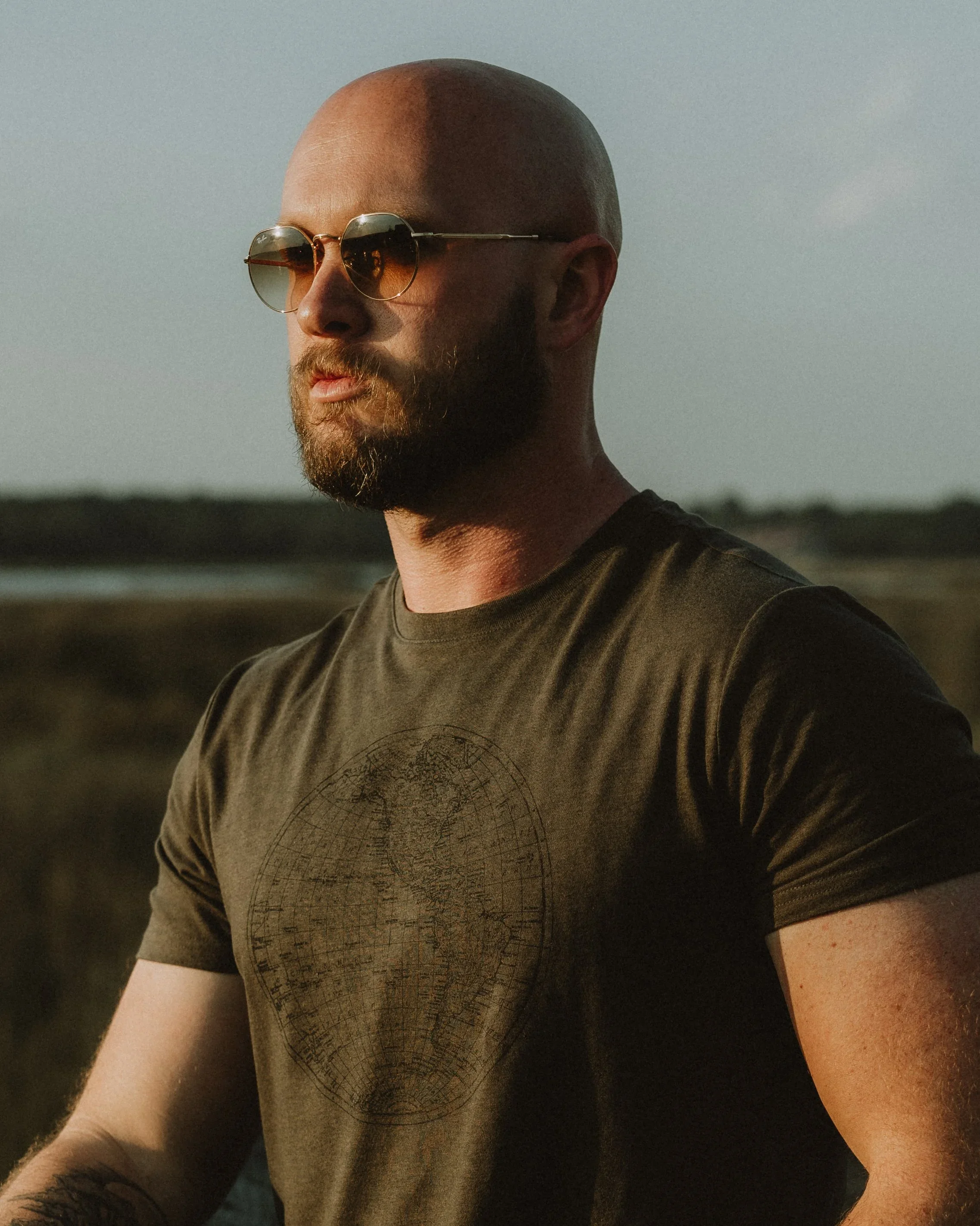A man with sunglasses, a beard, and a bald head, outdoors during sunset, wearing a dark T-shirt with a world map design.