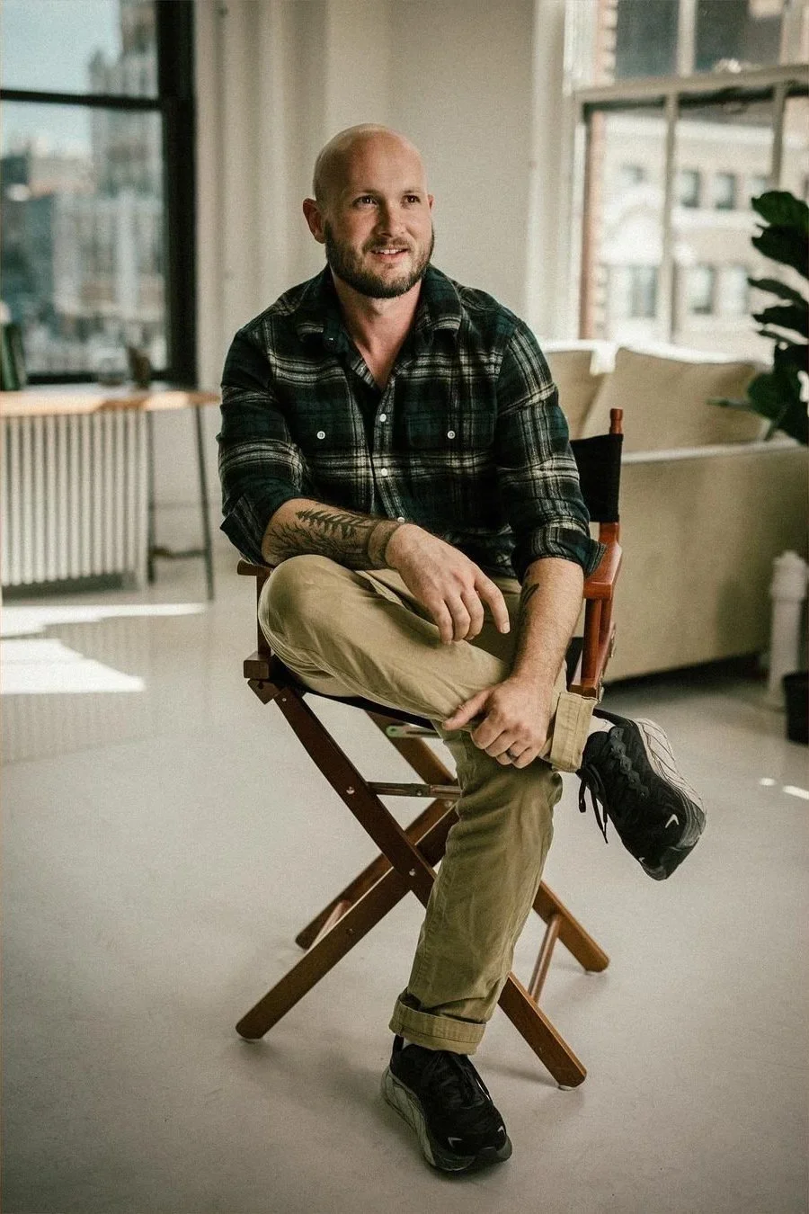 A man with a beard and tattoos, wearing a dark plaid shirt and khaki pants, sitting on a director's chair inside a modern apartment with large windows and city views.