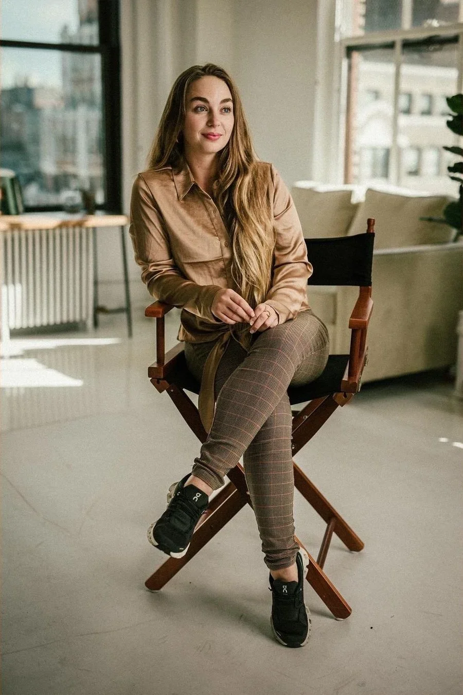 A young woman with long blonde hair sitting on a director's chair in a modern, well-lit apartment. She is wearing a tan button-up shirt, patterned jogger pants, and black sneakers, with a calm and confident expression.
