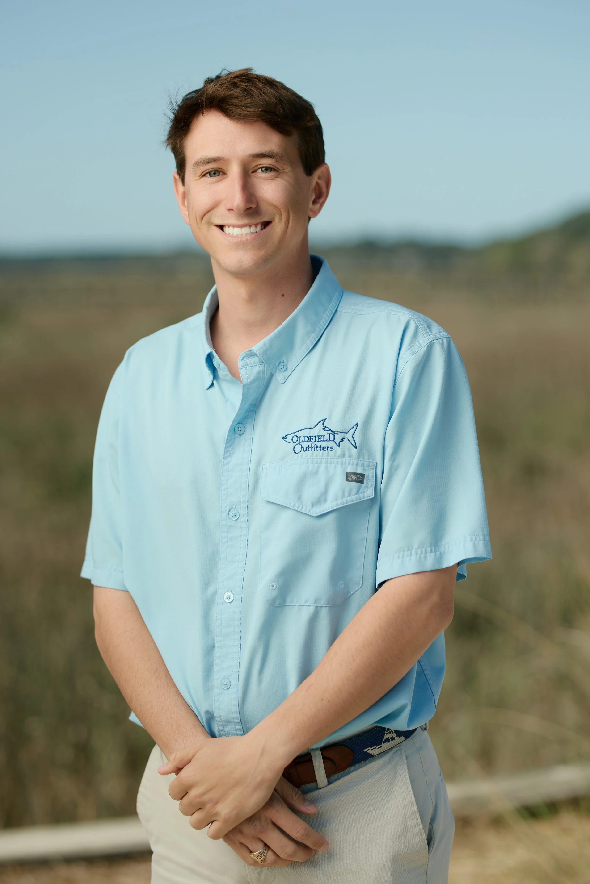 A young man smiling outdoors, wearing a light blue button-up shirt with a logo that reads 'Oldfield Outfitters' and a fish illustration, standing in a grassy field with a blurred natural landscape background.