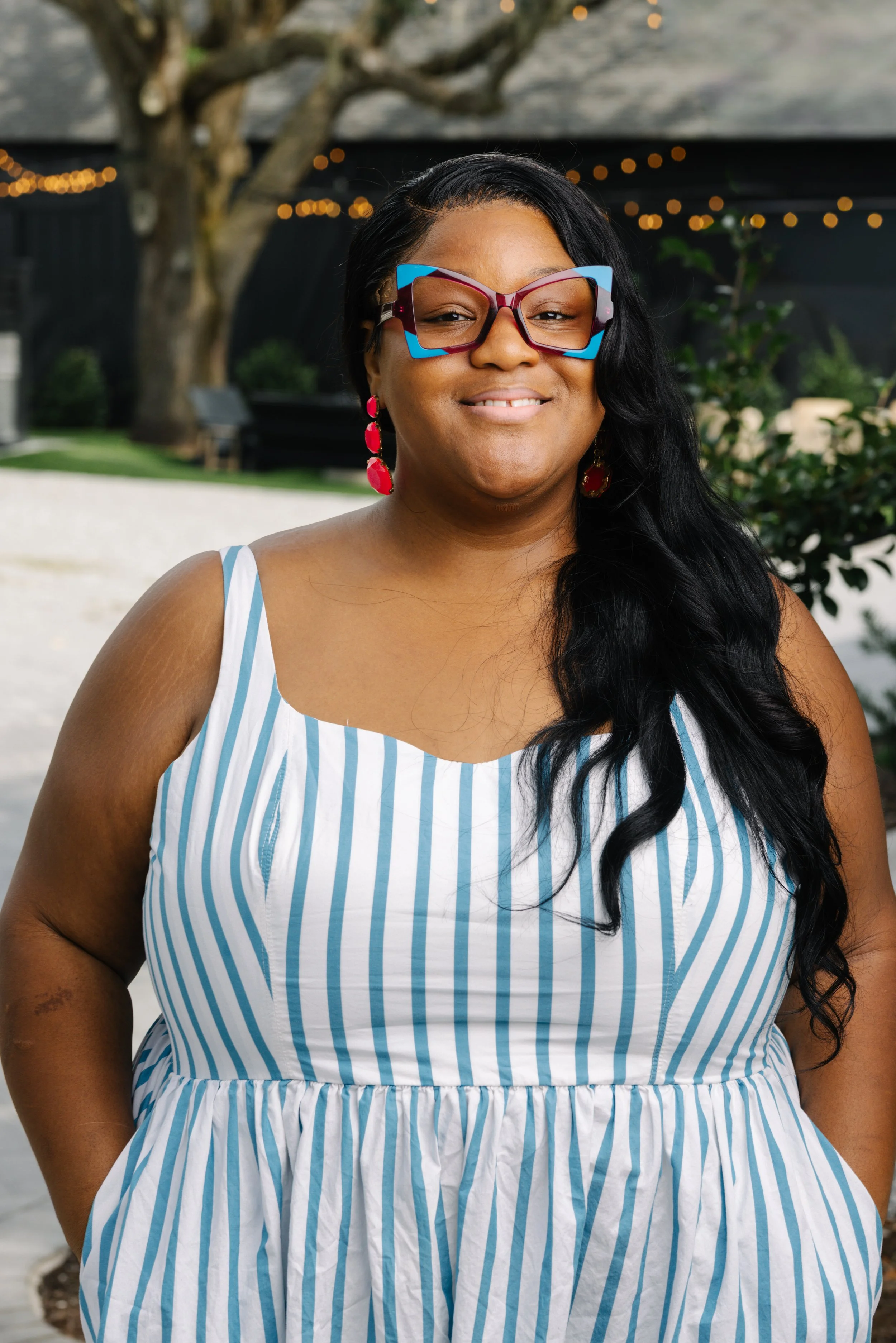A woman smiling outdoors with dark, wavy hair wearing colorful sunglasses, pink earrings, and a blue and white striped sleeveless dress.