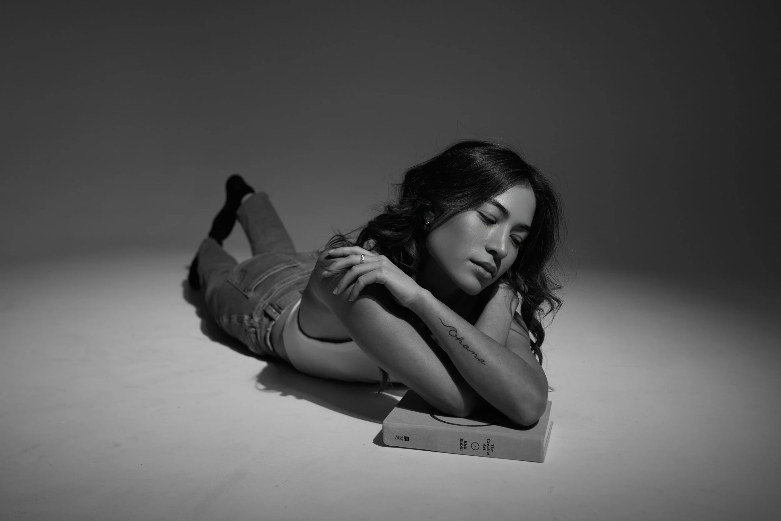 A woman with long dark hair lying on her stomach on a white surface, resting her head and arms on a closed book, with her eyes closed and a calm expression in a black-and-white photo.