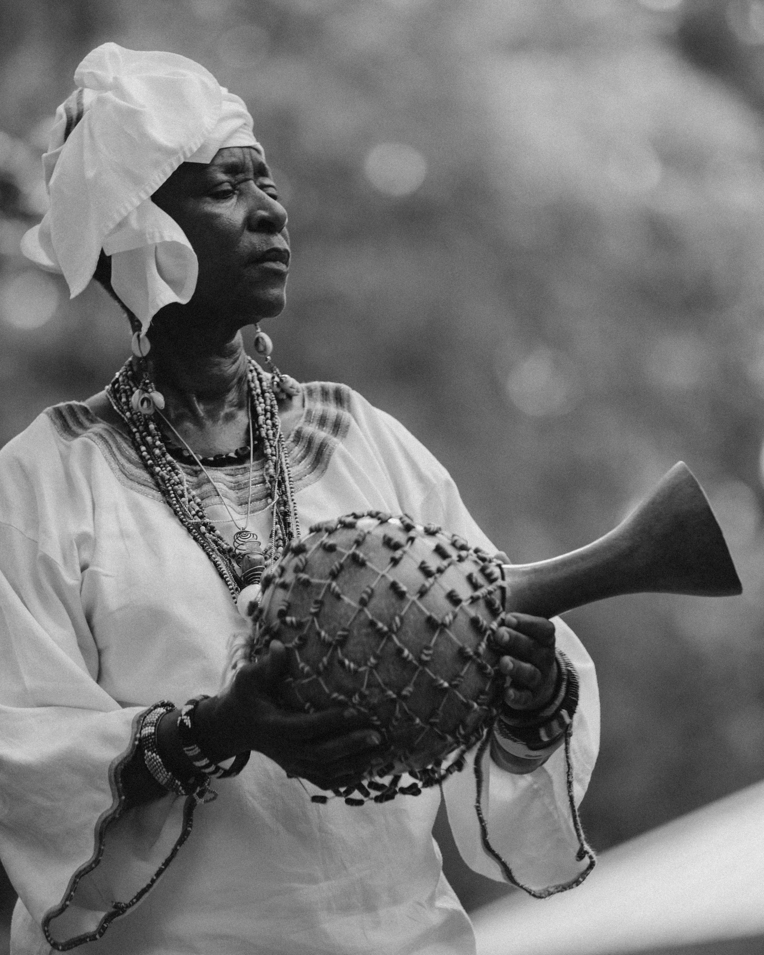 An elderly woman wearing traditional attire and jewelry, holding a decorated drum with a horn in her hand, against a blurred background.