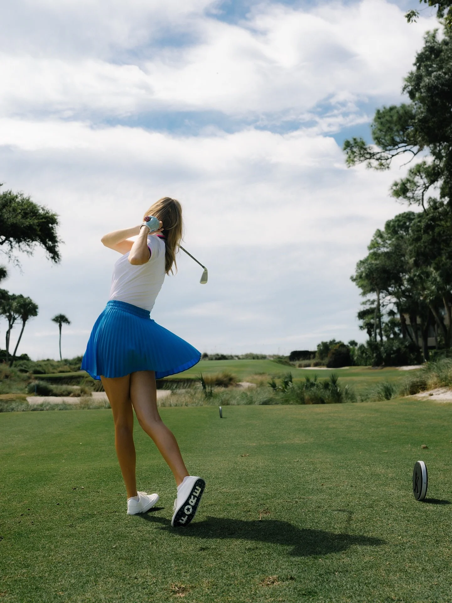 A woman in a white shirt and a blue pleated skirt swings a golf club on a golf course under a partly cloudy sky, with trees and green landscape in the background.