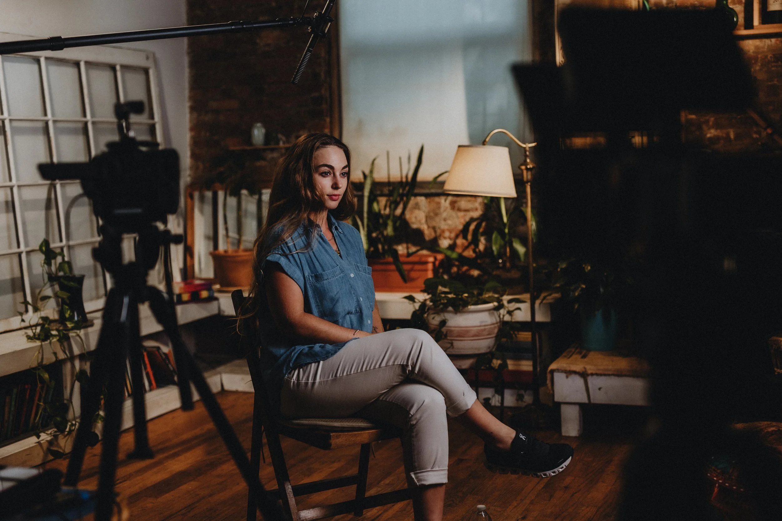 A woman seated on a chair during a video shoot or interview, surrounded by plants and warm lighting in a cozy indoors setting, with a camera, microphone, and studio lights set up.