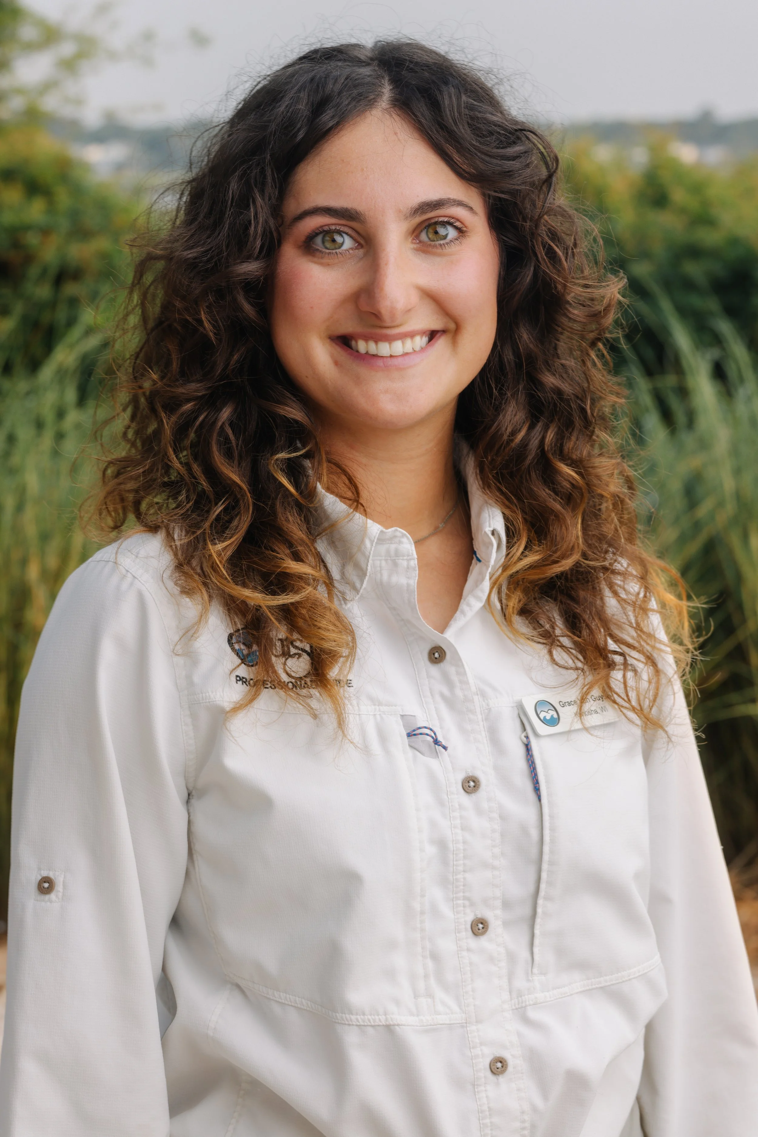 A woman with long, curly brown hair and light green eyes smiling outdoors. She is wearing a white collared shirt with a name tag. Background features blurred green foliage.