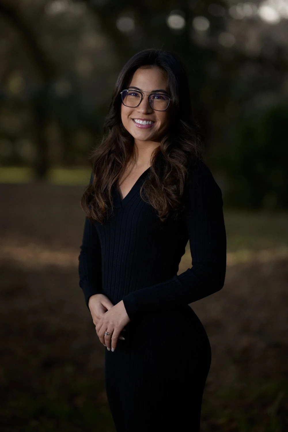 A woman with long dark hair, wearing glasses and a black dress, standing outdoors in a park at dusk, smiling at the camera.