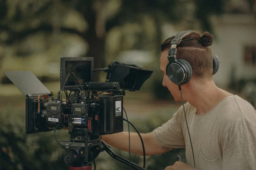 Filmmaker or videographer operating a professional camera outdoors with headphones on, focusing on capturing footage in a park-like setting.