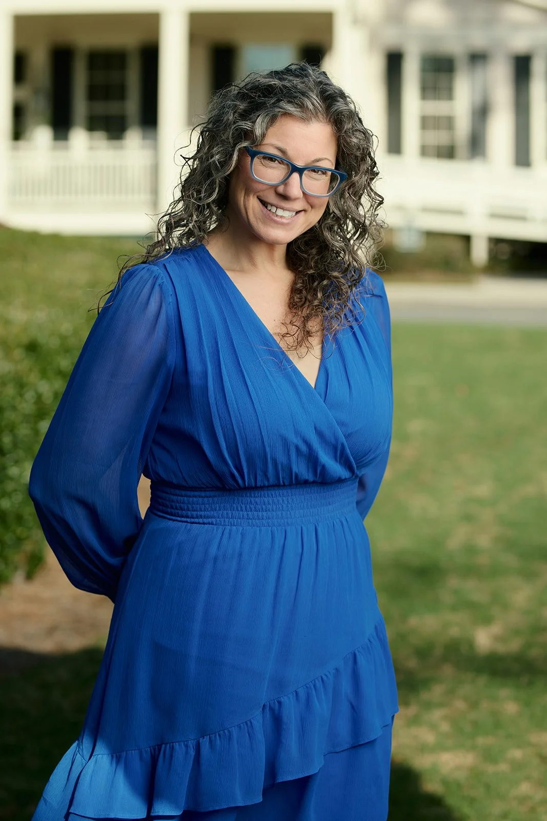 A smiling woman with curly gray hair and blue glasses standing outdoors in front of a house, wearing a long blue dress with pleats and ruffles.