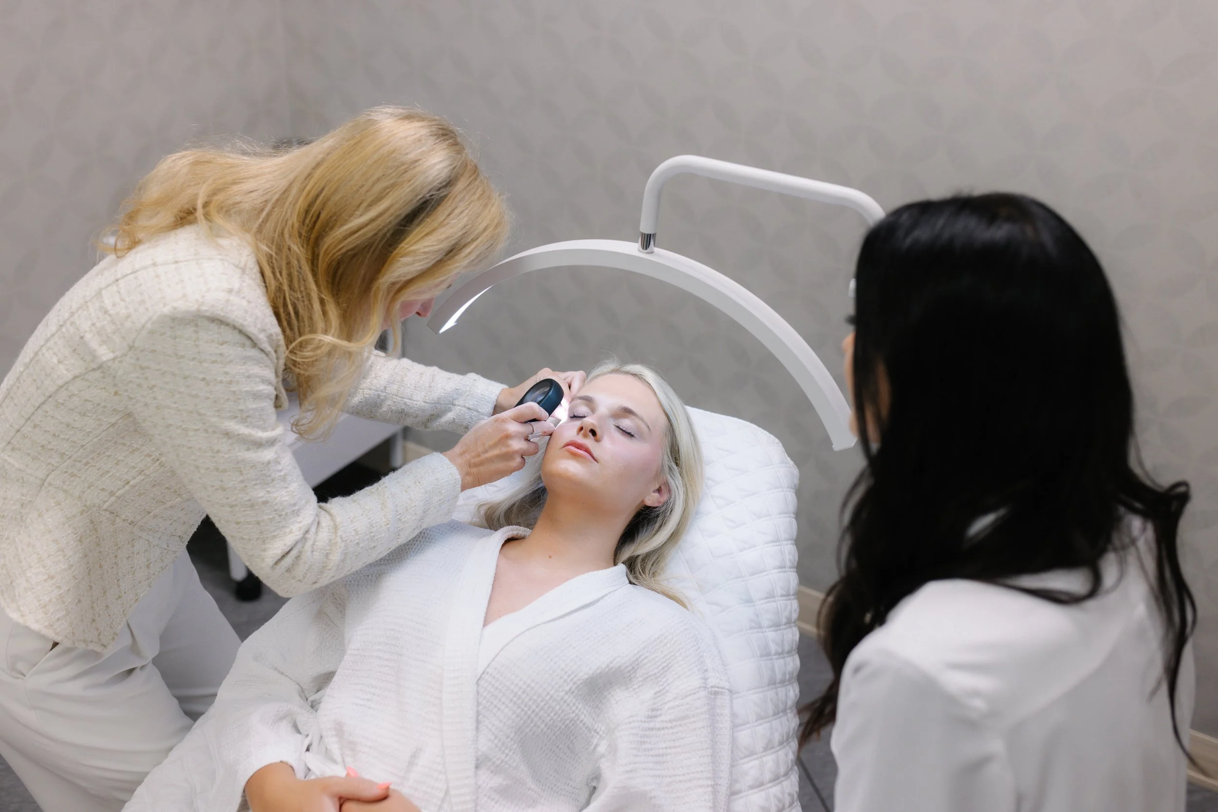 A woman lying on a treatment bed with a white quilted pillow, receiving a facial skincare treatment using a handheld device from a professional woman with blonde hair, while another professional woman with dark hair observes.