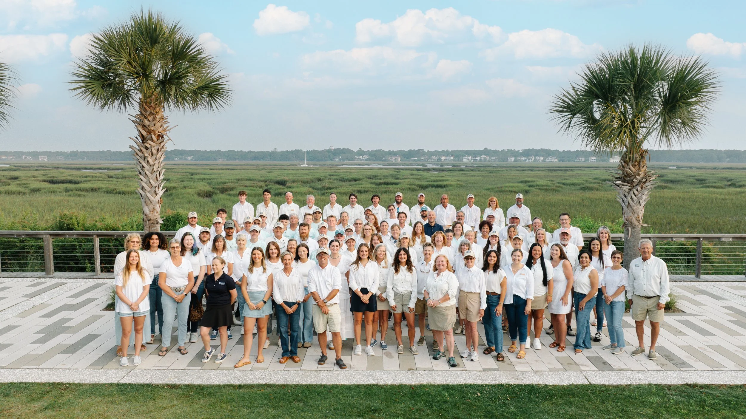 Group of people standing outdoors on a tiled patio with palm trees and a marshland in the background.