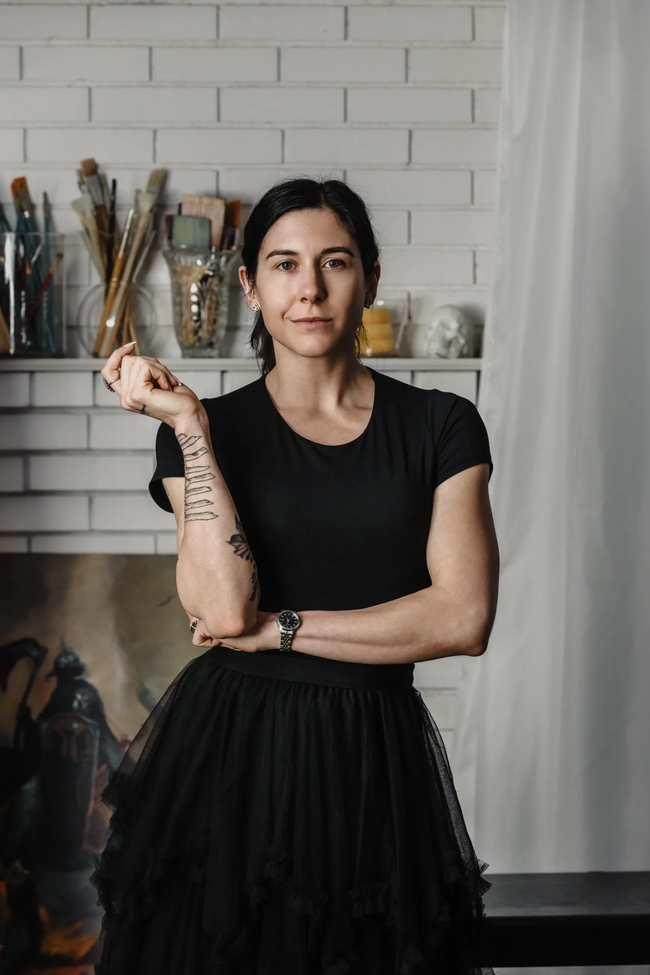 A woman with short black hair and tattoos on her left arm, wearing a black t-shirt and a black tulle skirt, standing in an art studio or gallery with paintbrushes and art supplies in the background.